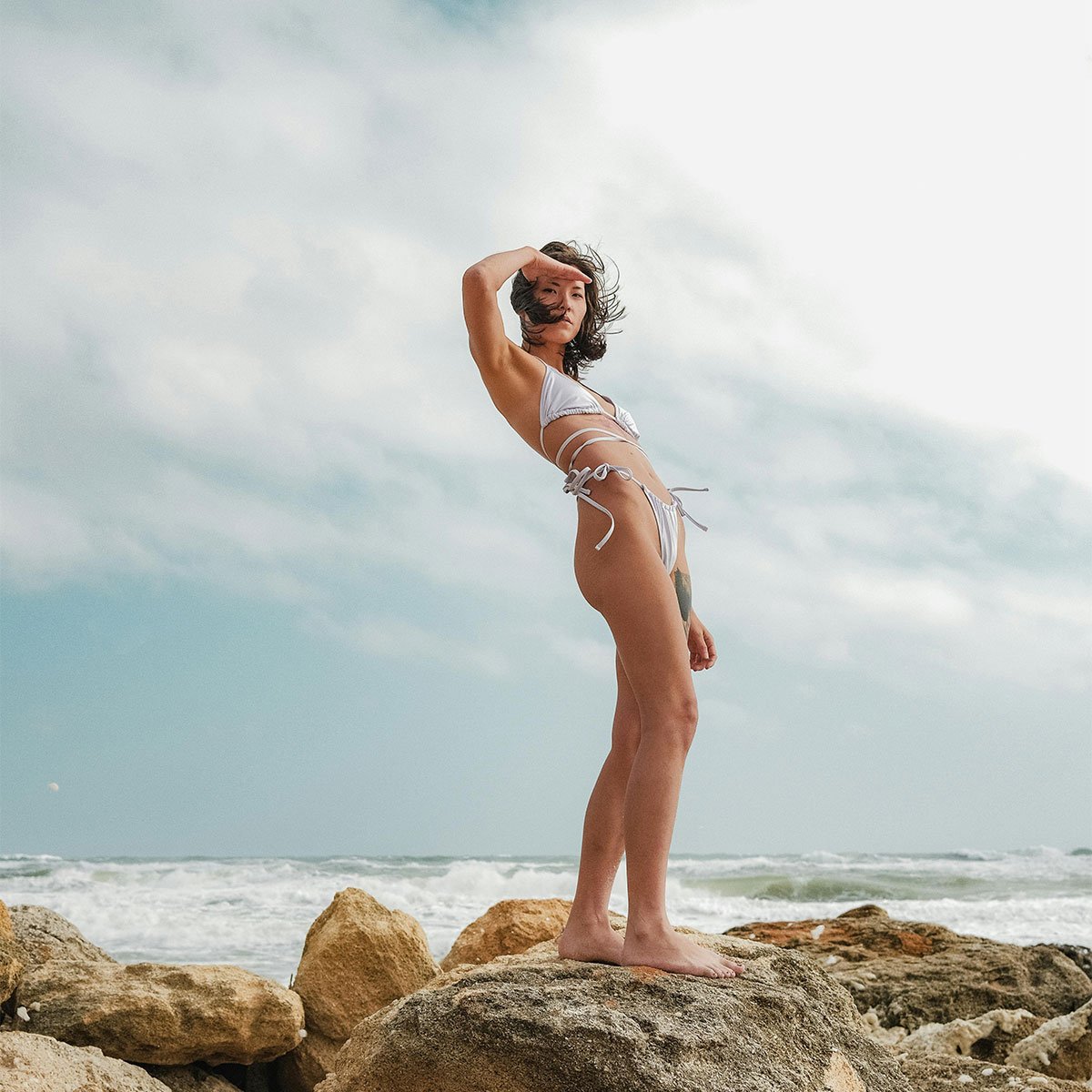 Woman in white bikini standing on seaside rocks under tropical sky, symbolizing freedom and confidence at Casa Amorsol.