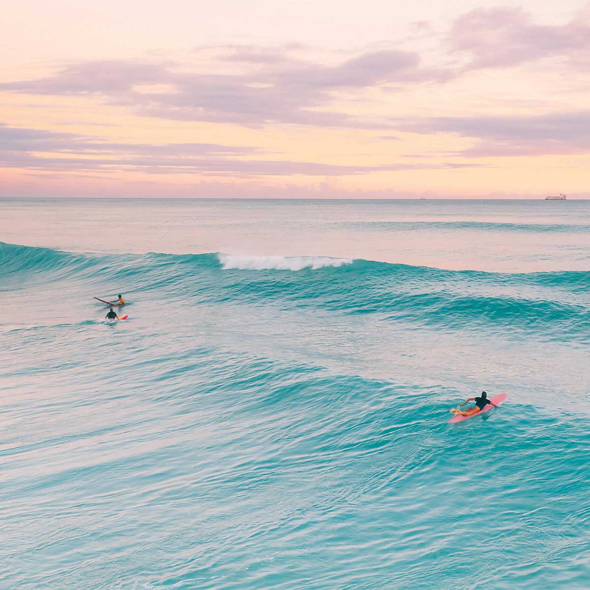 Surfers waiting on gentle ocean waves under a pastel sky — relaxed island rhythm.