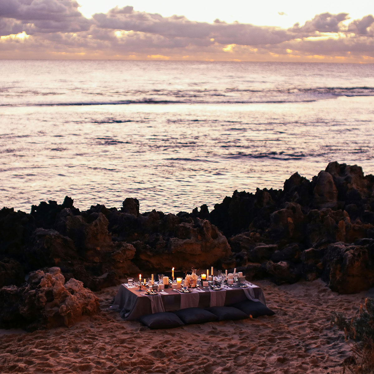 Romantic table setup with candles on the sand overlooking the sea at dusk.