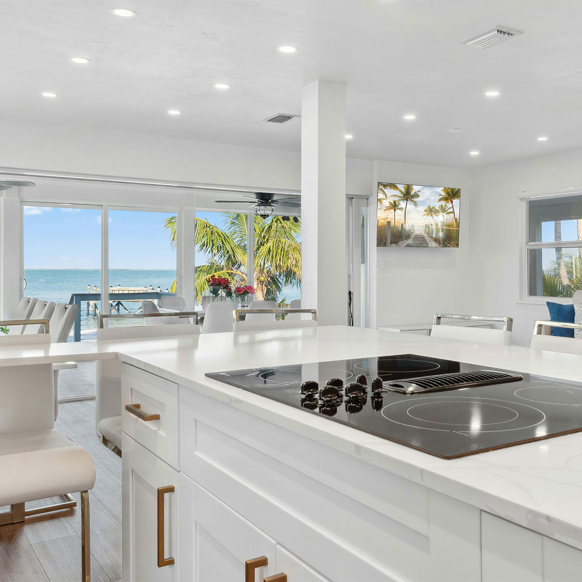 Open-concept kitchen at Casa Amorsol with white cabinetry, brass details, and panoramic ocean view through large glass windows overlooking palm trees and the beach.