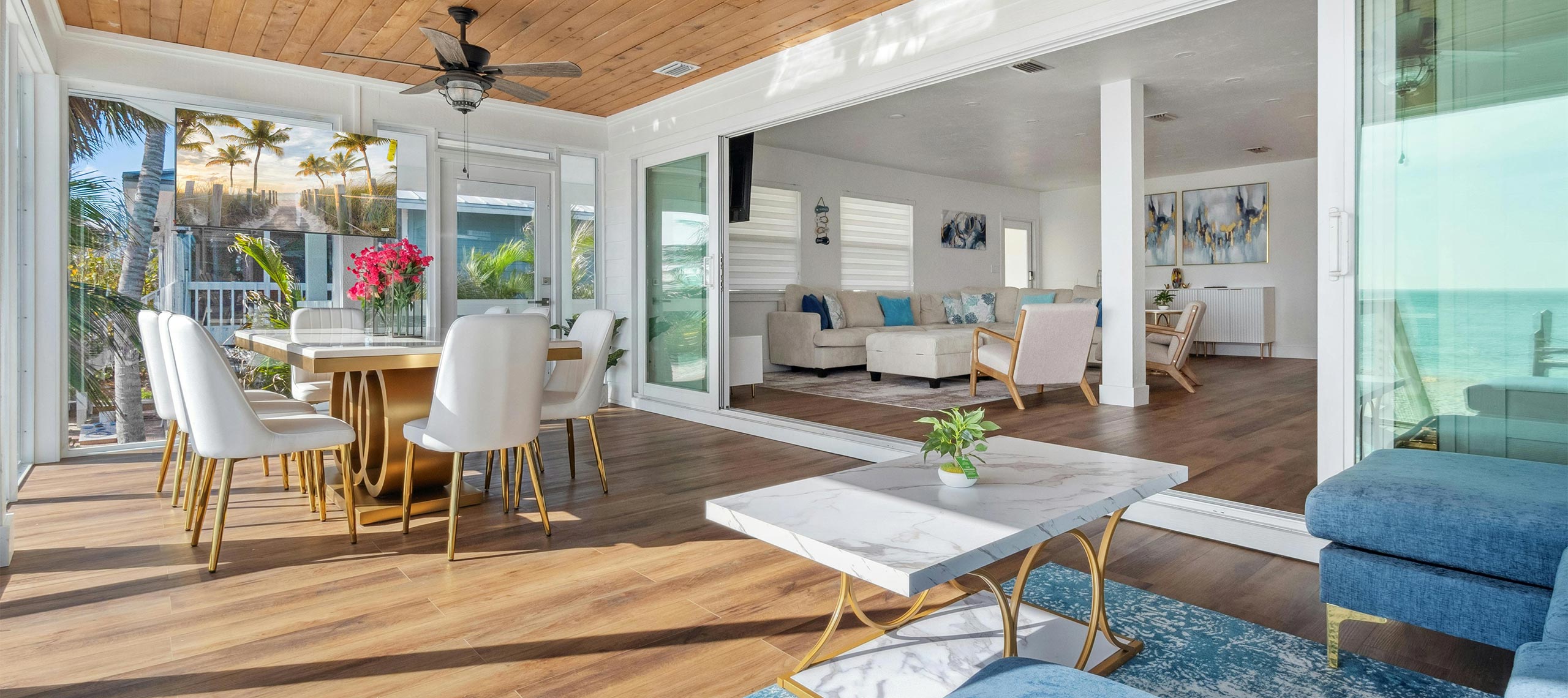 Bright open-concept living and dining area with ocean view, modern white chairs, wood ceiling, and natural sunlight streaming through glass doors — elegant beachfront villa interior.