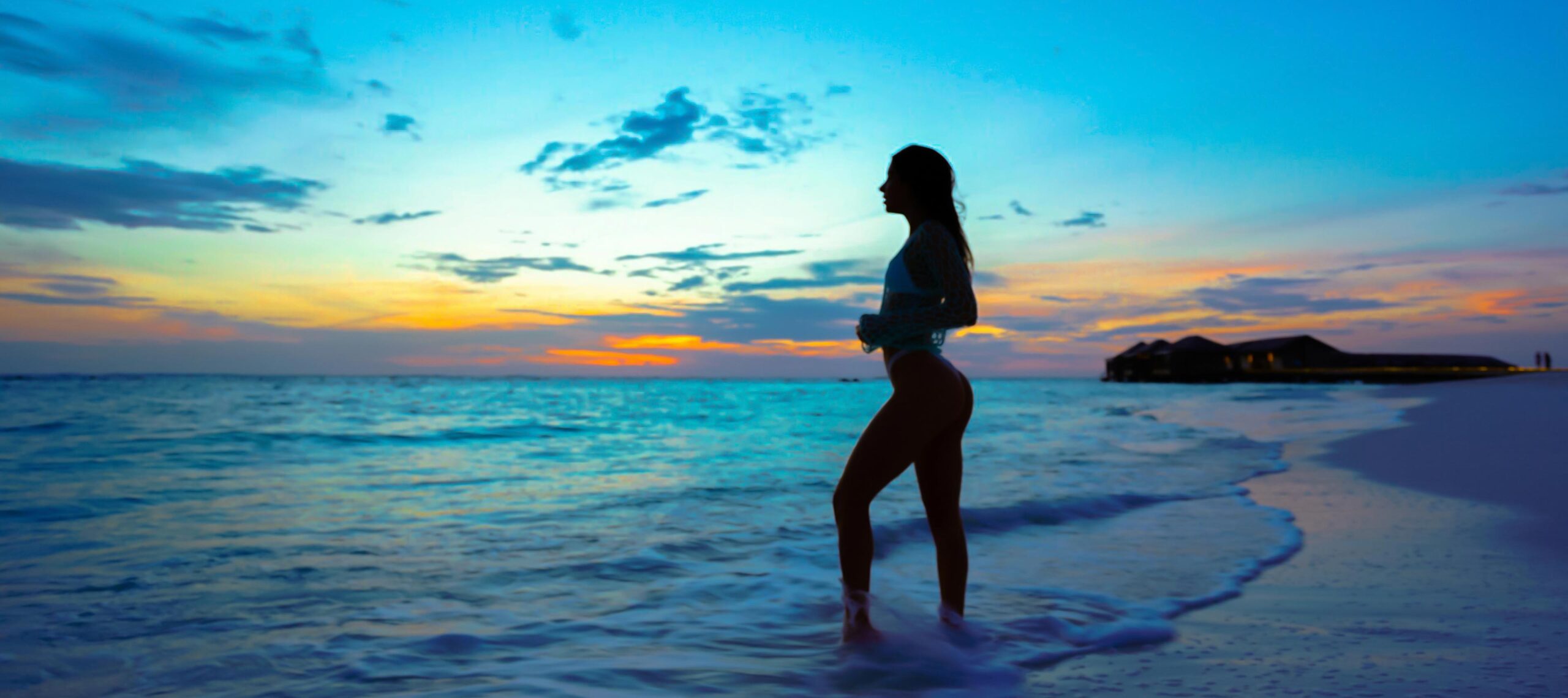 Silhouette of a woman standing at the shoreline during a colorful sunset, waves touching her feet near beachfront villas — evoking peace, freedom, and oceanfront living.